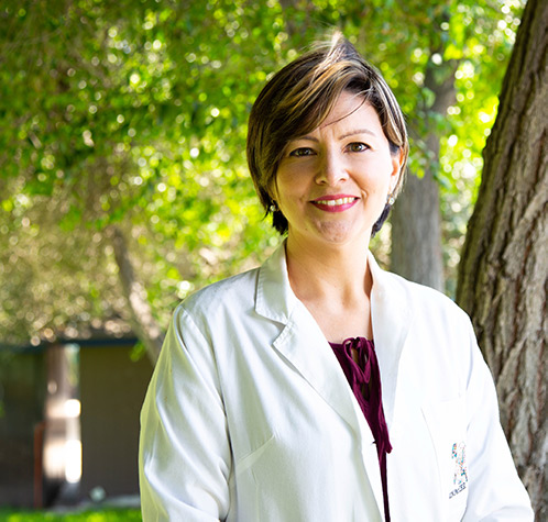 med-gov-staff Smiling woman in white medical coat, standing outside with trees in background